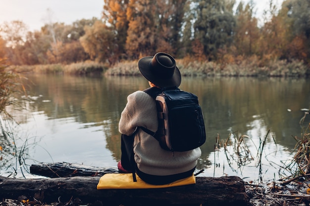 Traveler with backpack sitting on river bank at sunset. Middle-aged woman admiring autumn nature