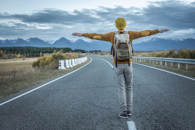 Photo traveler with a backpack in a yellow hat and a sweater on a road, against the background of mountains is standing with his arms outstretched to the sides. concept of freedom, autumn, travel, hiking