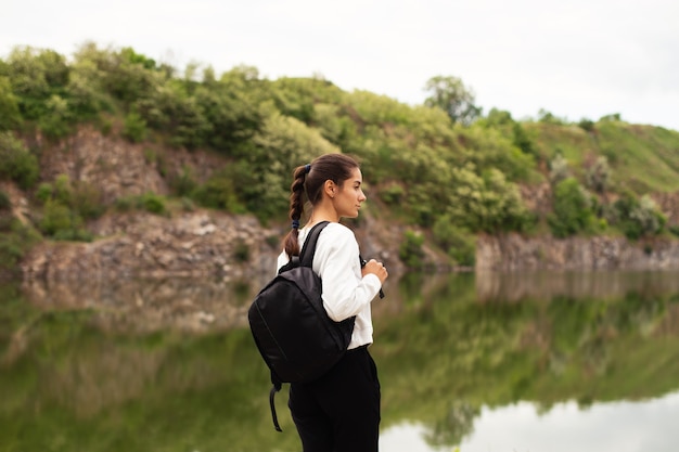 Traveler woman enjoys a beautiful view of the lake. Slender young woman travels with a backpack and a tripod.