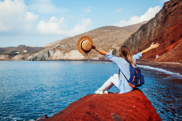 ギリシャ、サントリーニ島の赤いビーチの岩に腕を上げるリラックスした旅行者の女性。旅行と休暇
