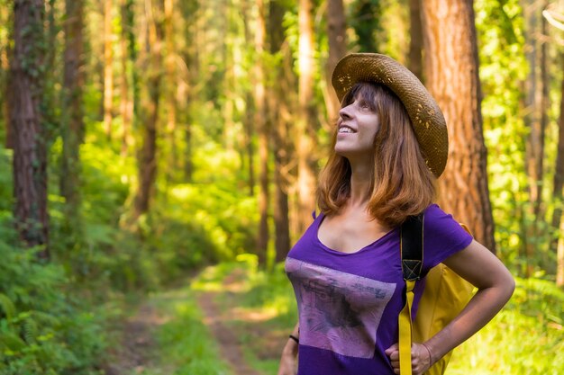 Женщина-путешественница улыбается в шляпе и смотрит на лесные сосны