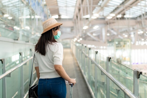 エスカレーターで空港に立ってターミナルの外を見てマスクを着て荷物を持つ旅行者女性