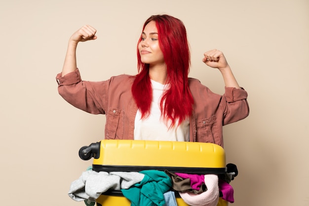 Traveler woman with a suitcase full of clothes isolated on beige wall doing strong gesture