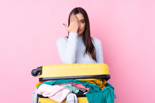 Traveler woman with a suitcase full of clothes over isolated pink wall covering a eye by hand