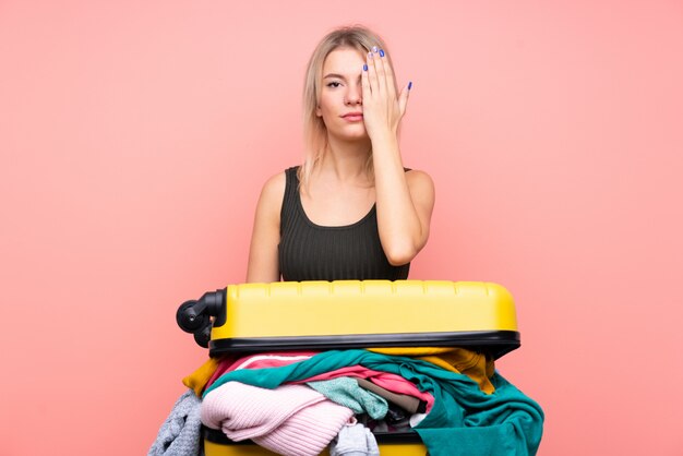 Traveler woman with a suitcase full of clothes over isolated pink wall covering a eye by hand