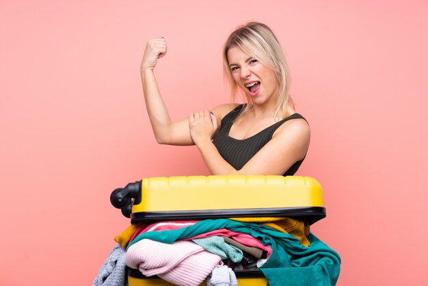 Traveler woman with a suitcase full of clothes over isolated pink wall making strong gesture