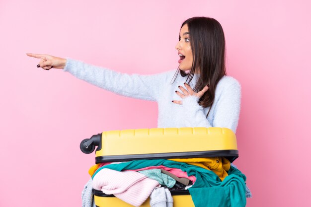 Traveler woman with a suitcase full of clothes over isolated pink wall pointing finger to the side