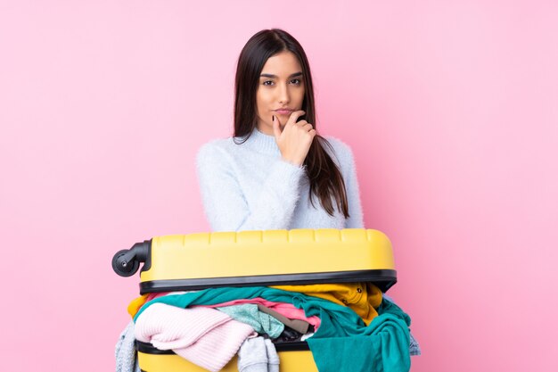 Traveler woman with a suitcase full of clothes over isolated pink wall thinking an idea