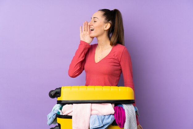 口を大きく開けて叫んで分離の紫色の壁の上の服の完全なスーツケースを持つ旅行者女性