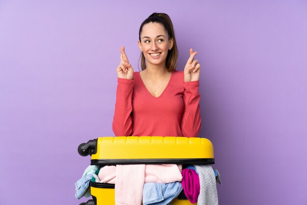 Traveler woman with a suitcase full of clothes over isolated purple wall with fingers crossing