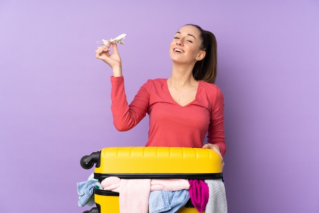 Traveler woman with a suitcase full of clothes over isolated purple wall