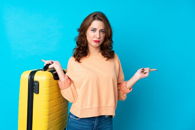 Traveler woman with suitcase over isolated blue  pointing to the laterals having doubts