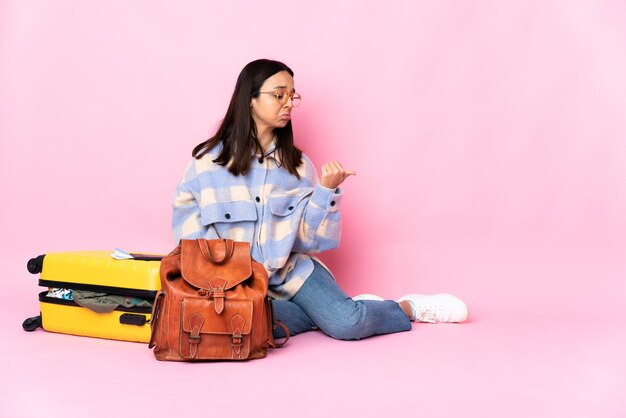 Traveler woman with a suitcase sitting on the floor unhappy and pointing to the side