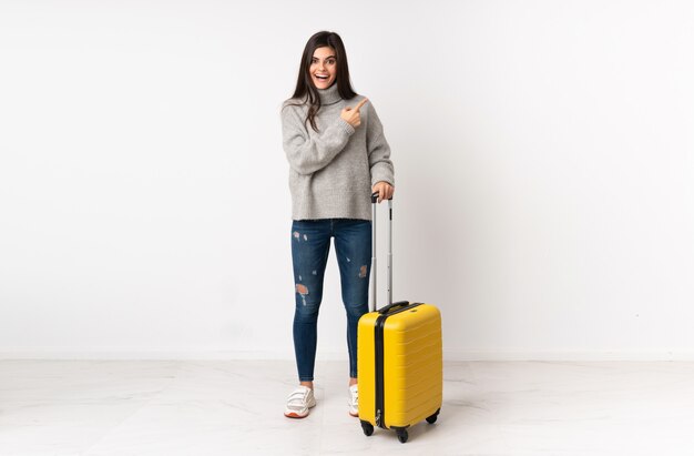 側に指を指している白い壁の上のスーツケースを持つ旅行者女性
