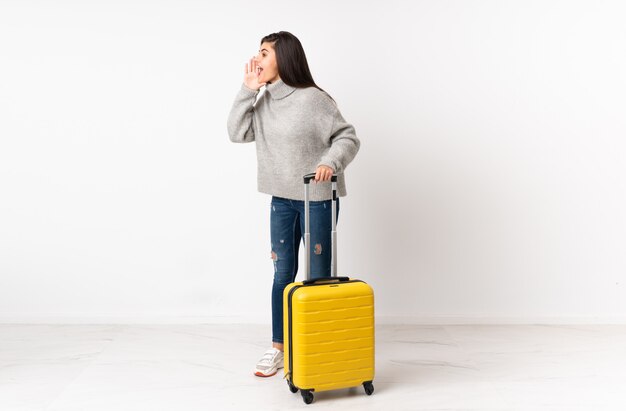 traveler woman with a suitcase over white wall shouting with mouth wide open