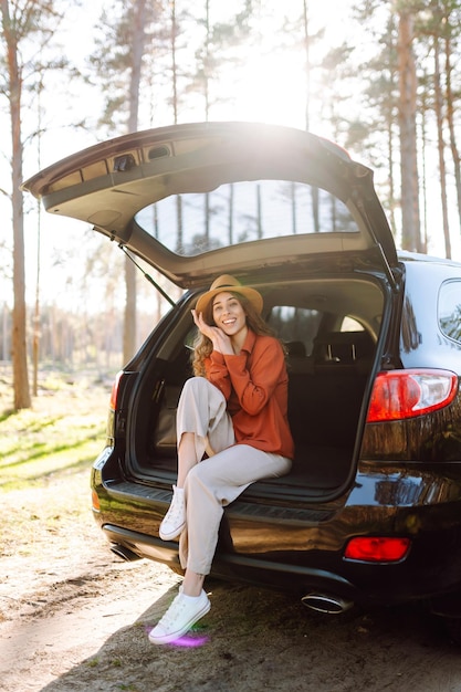 a traveler young woman smiles while sitting in the trunk of her car during a sunny day in a forest