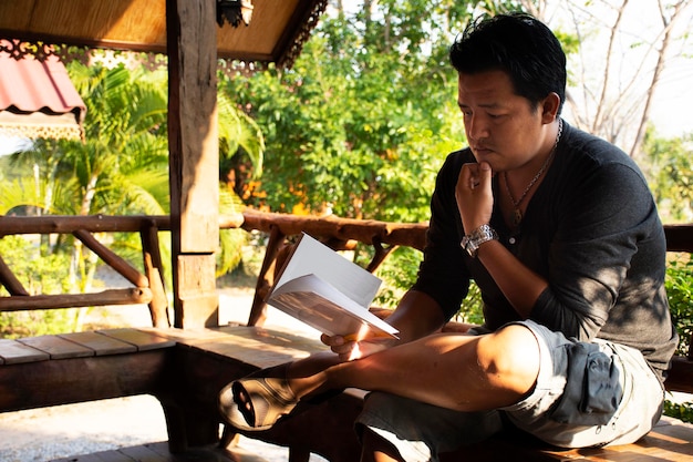 Travelers thai man sitting relaxed reading book at wooden hut\
of resort and homestay after rent and rest in morning time at the\
city of phraya lae the brave in chaiyaphum thailand