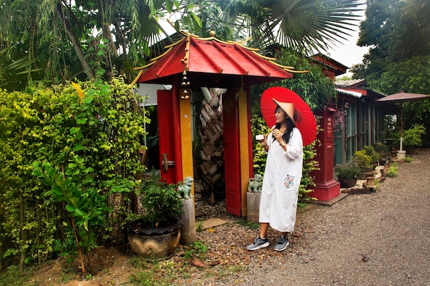 竹の帽子と赤い傘で服を着ている旅行者のタイの女性は、ラチャブリタイの朝の時間にリゾートホテルの庭とヴィンテージのレトロな家で写真を撮るために