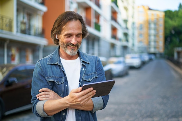 Traveling handsome mature man looking at digital tablet in hand talking outdoors having a call on urban city streets Mature man listening music use wireless earphones while travel old town streets