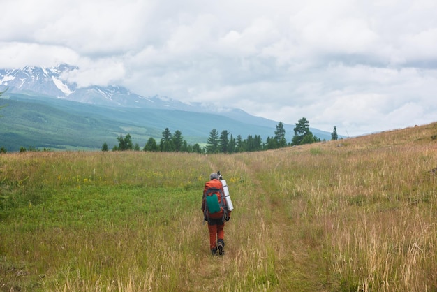 Traveling man in red with big backpack on way to snowy mountain range in low clouds Backpacker walks through forest to large mountains under cloudy sky Dramatic landscape with tourist in mountains