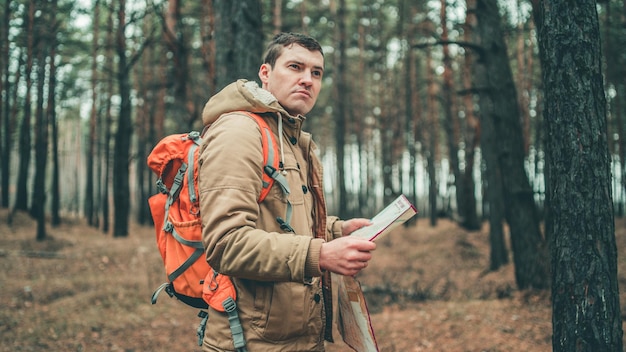 森の中の地図を持つ旅行者寒い天気で木の近くに立っているバックパックを持つハンサムな男の肖像画