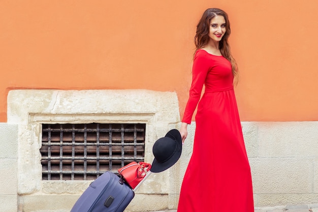 Путешествующая женщина в красном длинном платье тянет чемодан на улице города.