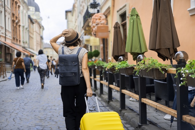 Foto turista della donna in viaggio in cappello con zaino e valigia camminando lungo la strada