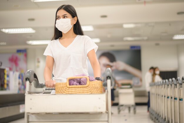 A traveling woman wearing protective mask in the airport with luggage on trolley, travel under Covid-19 pandemic, safety travels, social distancing protocol, New normal travel concept