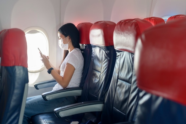 A traveling woman wearing protective mask onboard in the aircraft using smartphone, travel under Covid-19 pandemic