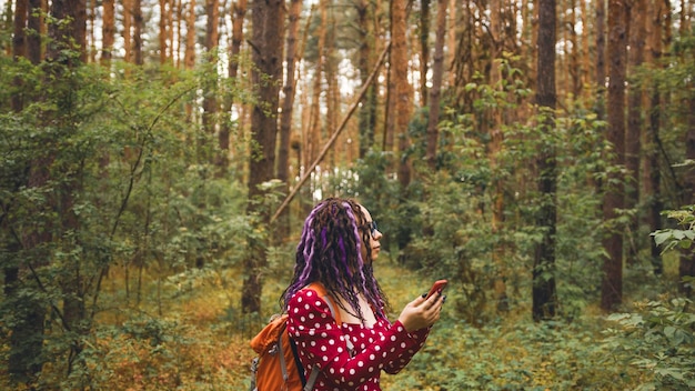 Traveling woman with dreadlocks with a phone in woods Female traveler with backpack in woods reading map on a smartphone and looking for a way in cloudy weather