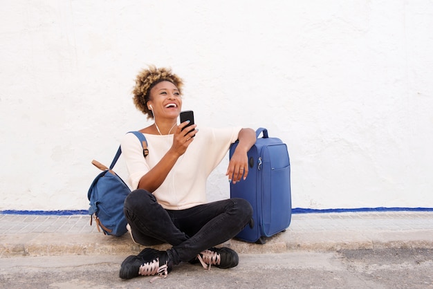 Traveling young woman sitting on sidewalk with suitcase and listening to music