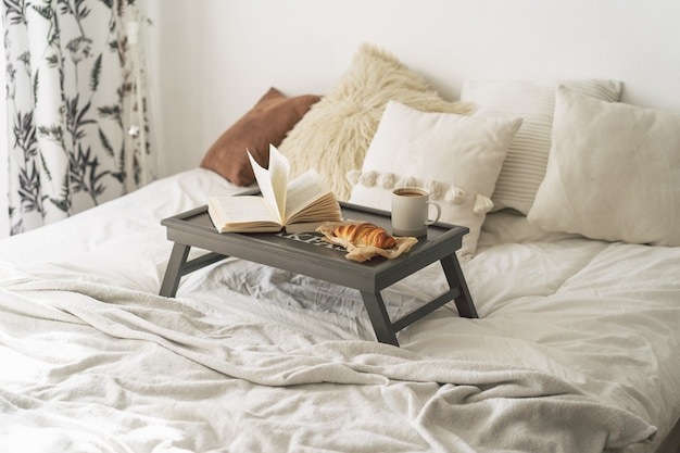 Tray with croissant, cup of coffee and book on white clean bed.