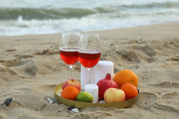 Tray with glasses of wine, fruits and candles on sandy sea beach