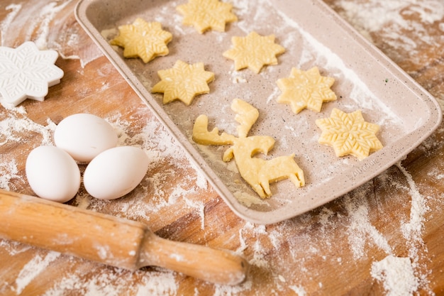 テーブルの近くに3つの卵、めん棒、小麦粉、カッターが付いた2列の生のクリスマスクッキーが入ったトレイ