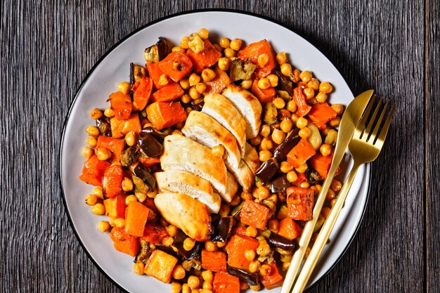 Traybake salad of chickpea with butternut, eggplant, with lemon and baked chicken breast, served on a plate with cutlery on a dark wooden table, top view, close-up