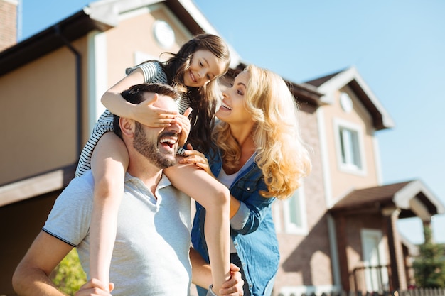 Photo treasured moments. cute little girl sitting on the shoulders of her father, covering his eyes with her hands while her mother standing near them and laughing