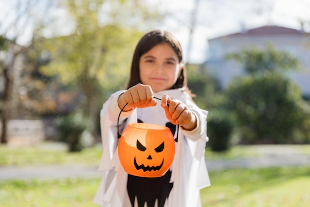 彼女の手にカボチャのバスケットを持って公園に立っているハロウィーンのための幽霊のカーニバルの衣装で笑顔の少女の屋外ショットを治療またはトリック