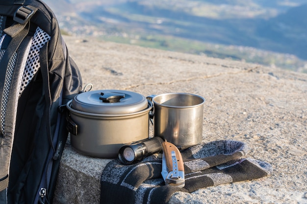 Trecking or hiking equipment set on concrete fence. Bagpack, socks, metal cup, kettle pot, folding knife, and flashlight. Outdoor activity concept.