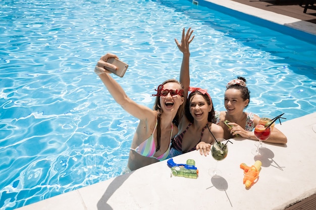 Три стильные улыбающиеся молодые девушки в купальниках делают салфи с коктейлями в бассейне на открытом воздухе в солнечный летний день