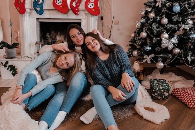 Tree young woman girls sitting next to the fireplace and christmas tree. having fun, togetherness and friendship. girls power. new year celebration. cozy winter days. home sweet home.