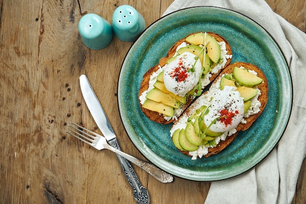 Trending food - avocado toast with ricotta, poached egg on rye bread in a ceramic plate on a wooden surface. Healthy breakfast meal
