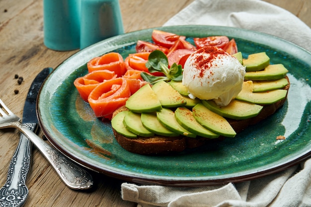 Trending food - avocadotoost met gepocheerd ei op roggebrood met zijdeachtige zalm, tomaat in een keramische plaat op een houten oppervlak. Gezonde ontbijtmaaltijd