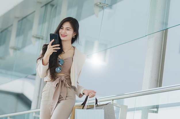 Trendy beautiful young Asian woman carrying colorful bags shopping online with mobile phone with copy space