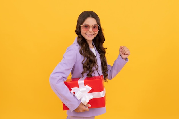 선글라스를 쓴 세련된 10대 소녀가 노란색 배경 쇼핑 할인에 선물 상자를 들고 있습니다.