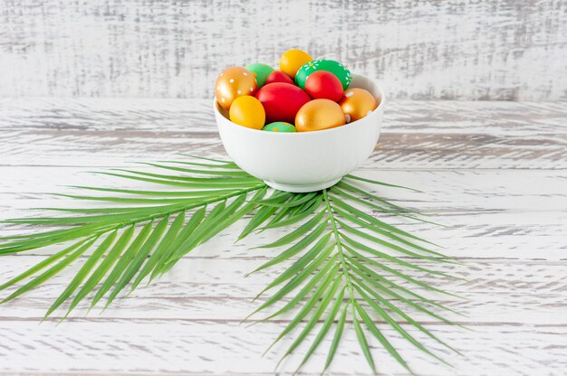 Trendy colored easter eggs, green tropical leaves and feathers on a plate on wooden table. Preparation for easter holidays. Fesstive composition