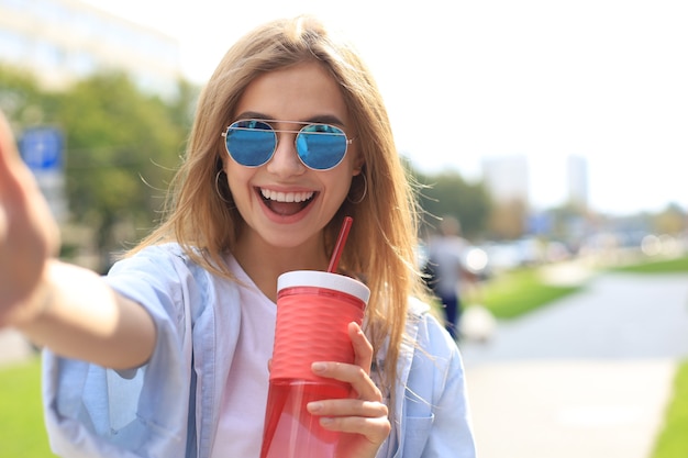 トレンディなクールな流行に敏感な女の子は、都会の街の背景でカクテルを飲みます。