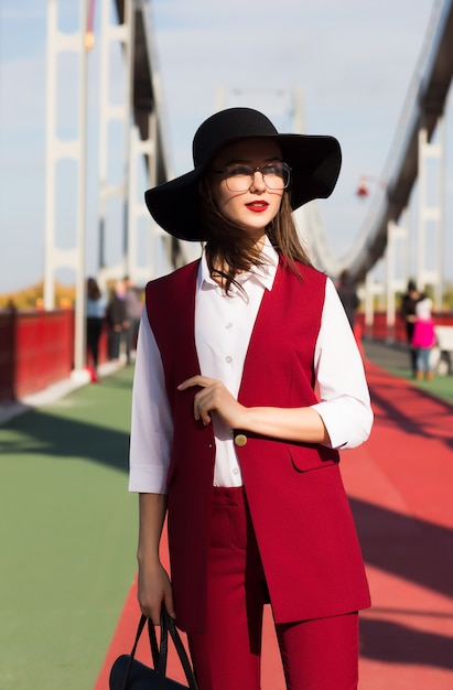 Trendy dressed brunette woman in red pantsuit and black hat posing at the bridge in sunny day