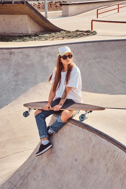 Trendy dressed young skater girl with longboard sitting on a skatepark.