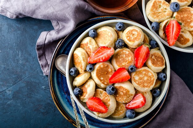 Trendy eten - pannenkoekengranen. Mini granen pannenkoeken met aardbeien en bosbessen in boul op blauwe cement achtergrond, bovenaanzicht. Kopieer ruimte over voor tekst.