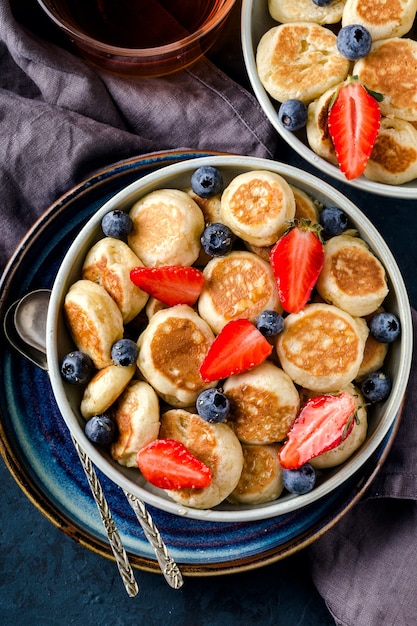 Trendy eten - pannenkoekengranen. Mini granen pannenkoeken met aardbeien en bosbessen in boul op blauwe cement achtergrond, bovenaanzicht. Verticaal formaat